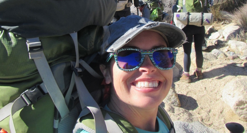 a veteran wearing sunglasses smiles at the camera while wearing a large backpacking